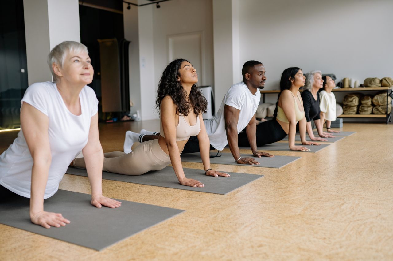 who-we-are A group of adults performing yoga poses on mats indoors, emphasizing diversity and wellness.